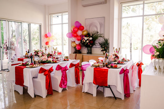 Brightly decorated indoor party with two long tables, white tablecloths, red runners, red and pink bows on chairs, floral centerpieces, and pink, red, and purple balloons. Large windows let in natural light. Prop hire, Prop hiring, Prop hire, event stylists Brisbane, brisbane event styling, brisbane event styling, event stylist Brisbane, Brisbane Wedding, Gold Coast Wedding, Floral Styling, Lush & Lavish Events