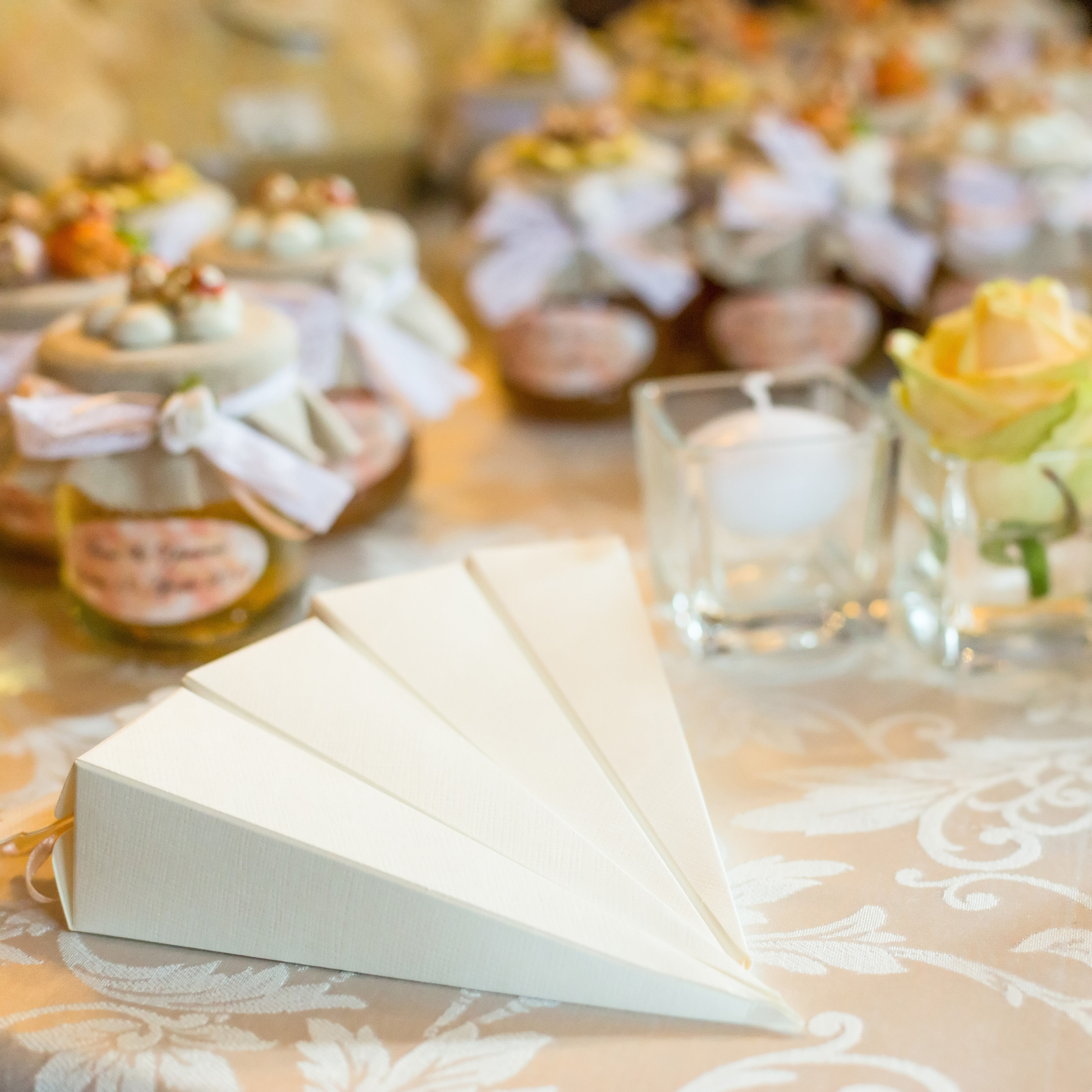 Decorated table with fabric-wrapped jars, a yellow rose in a vase, a candle, and triangular white boxes on a floral tablecloth. Prop hire, Prop hiring, Prop hire, event stylists Brisbane, brisbane event styling, brisbane event styling, event stylist Brisbane, Brisbane Wedding, Gold Coast Wedding, Floral Styling, Lush & Lavish Events, Brisbane Tablescape, Gold Coast Tablescapes, Brisbane Candle Hire