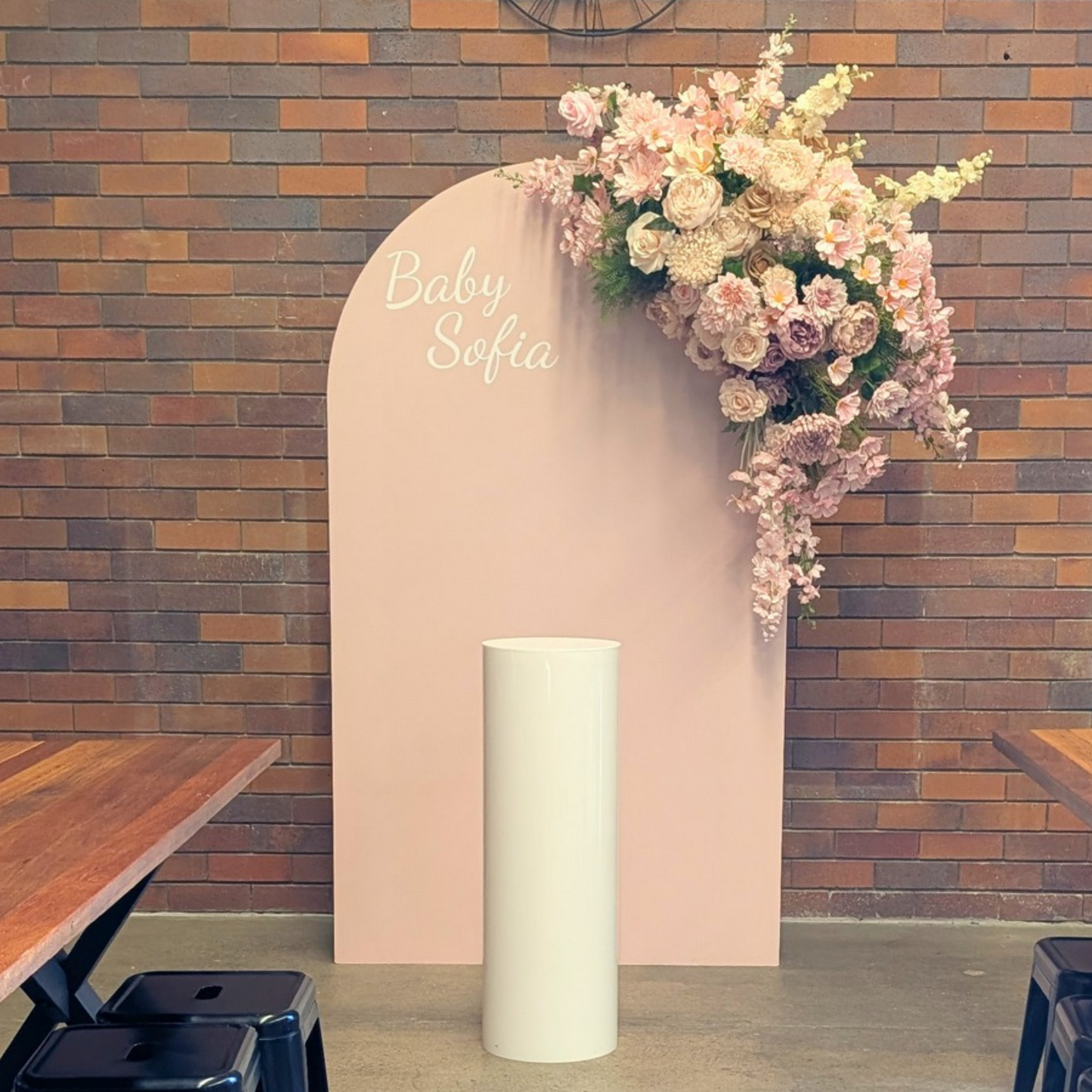 A baby shower backdrop setup featuring a tall, light pink arch with the text 'Baby Sofia' in white script, decorated with a cascading floral arrangement of pink and cream roses, set against a rustic brick wall with a white acrylic plinth. Styled at Future Brewing Woolloongabba by Lush and Lavish Events a Brisbane and Gold coast based Event styling, event planning, on-the-day coordination and Event MC services business.