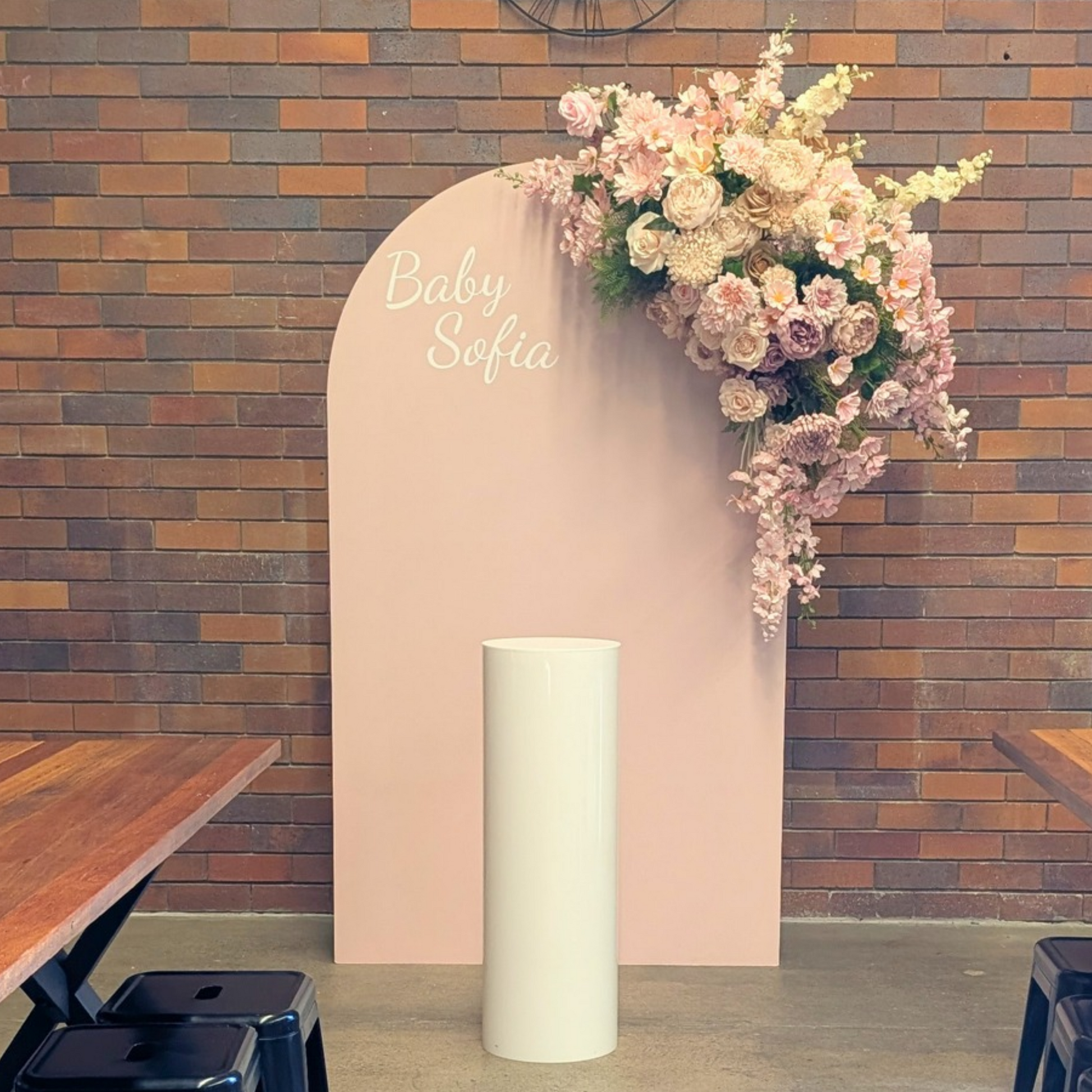 A baby shower backdrop setup featuring a tall, light pink arch with the text 'Baby Sofia' in white script, decorated with a cascading floral arrangement of pink and cream roses, set against a rustic brick wall with a white acrylic plinth. Styled at Future Brewing Woolloongabba by Lush and Lavish Events a Brisbane and Gold coast based Event styling, event planning, on-the-day coordination and Event MC services business.
