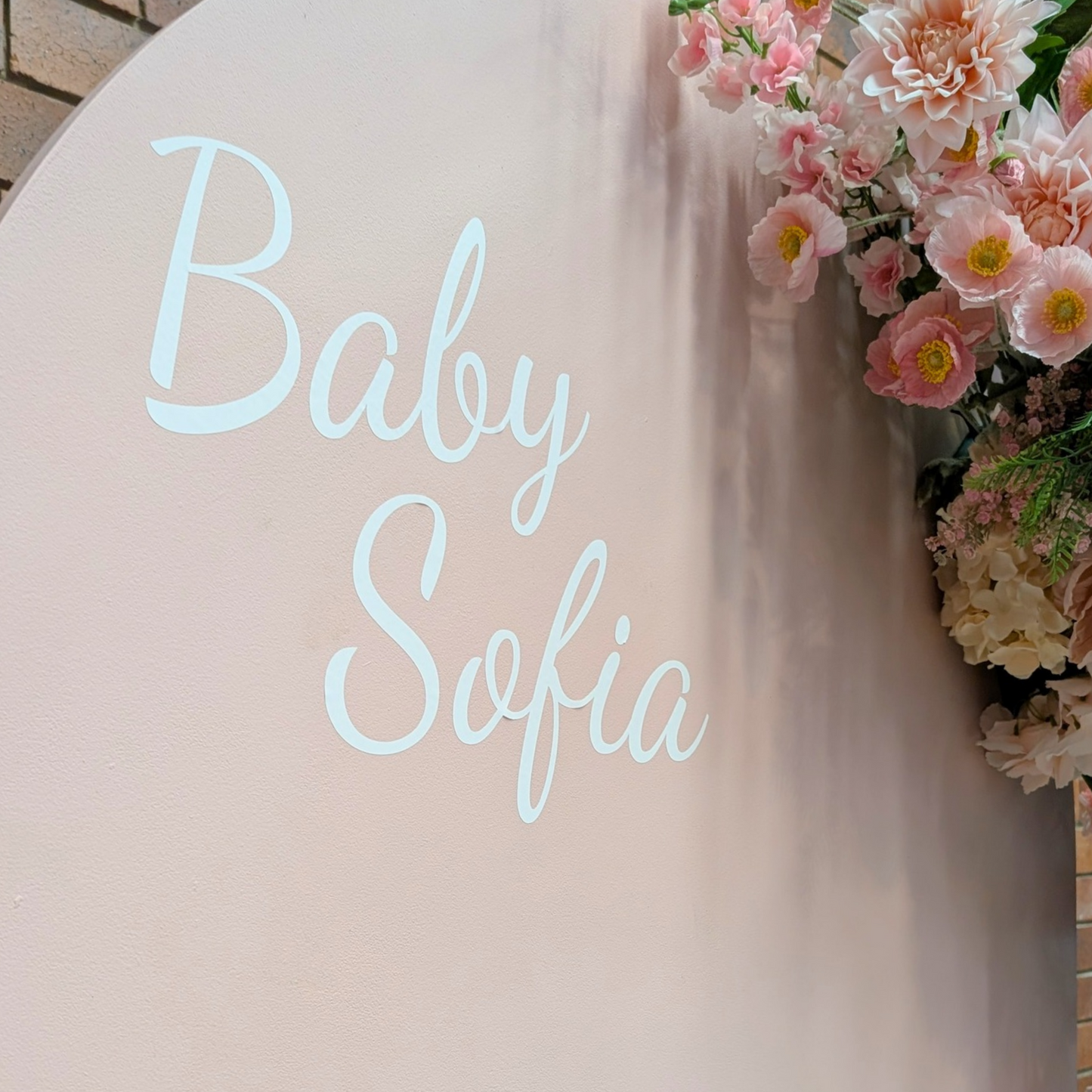 A baby shower backdrop setup featuring a tall, light pink arch with the text 'Baby Sofia' in white script, decorated with a cascading floral arrangement of pink and cream roses, set against a rustic brick wall with a white acrylic plinth. Styled at Future Brewing Woolloongabba by Lush and Lavish Events a Brisbane and Gold coast based Event styling, event planning, on-the-day coordination and Event MC services business.