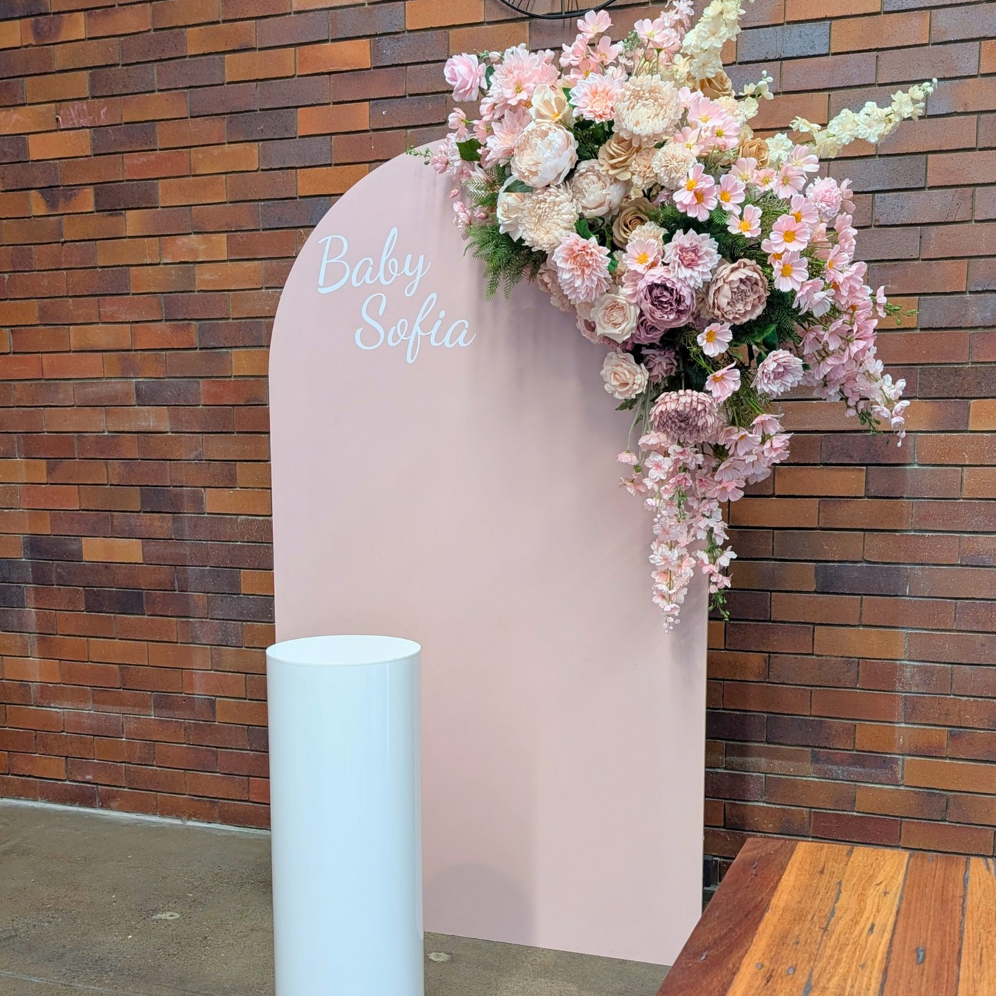 A baby shower backdrop setup featuring a tall, light pink arch with the text 'Baby Sofia' in white script, decorated with a cascading floral arrangement of pink and cream roses, set against a rustic brick wall with a white acrylic plinth. Styled at Future Brewing Woolloongabba by Lush and Lavish Events a Brisbane and Gold coast based Event styling, event planning, on-the-day coordination and Event MC services business.