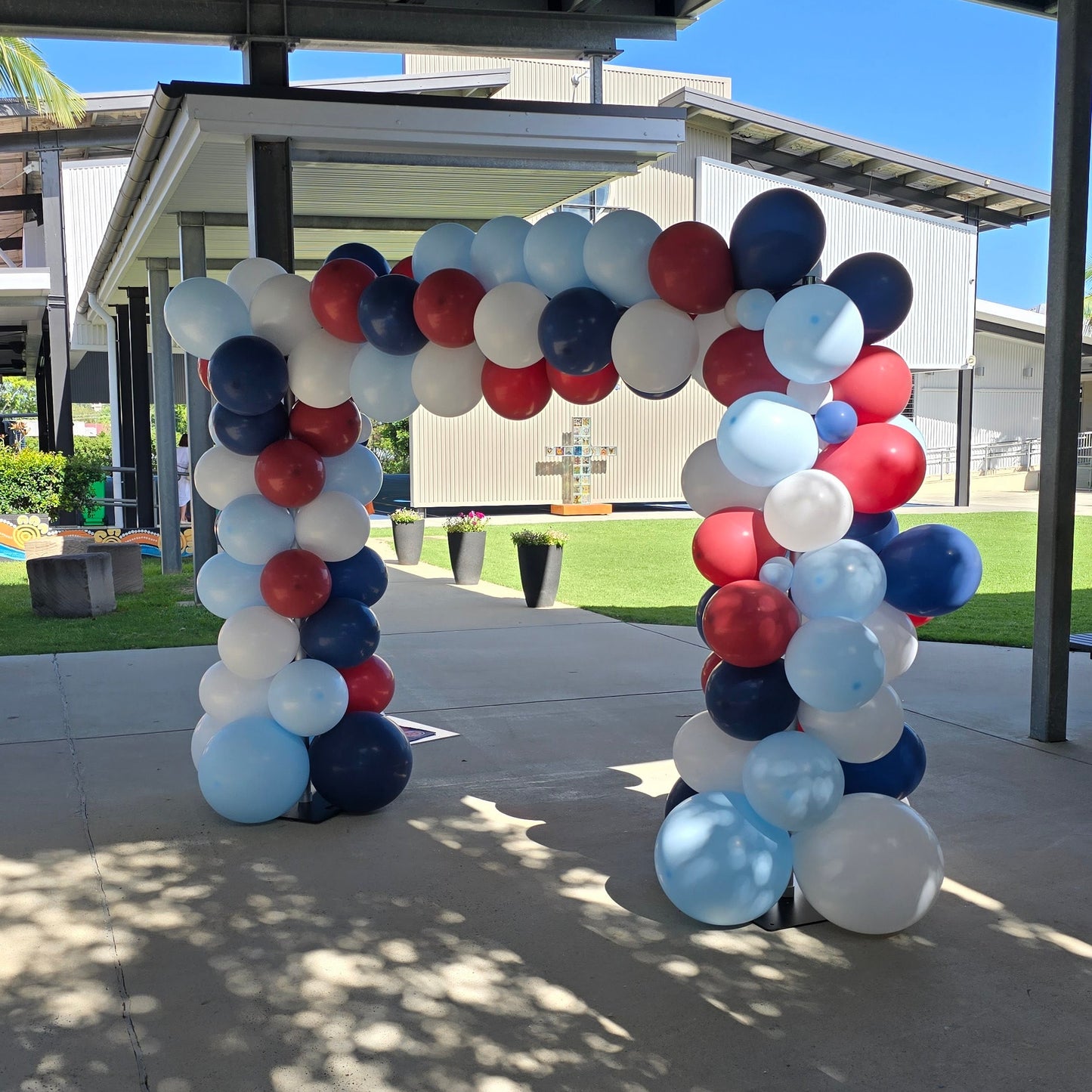 Balloon arch in red, white, and blue at an outdoor event under a covered area. organic balloon arch, corporate balloon arch, balloon arch Brisbane, balloon arch Gold Coast, corporate event styling Brisbane, brand activation balloons, product launch balloons, media wall balloon arch, event backdrop Brisbane, event backdrop Gold Coast, branded balloon arch, PR event balloons, company event decorations, business event balloons, balloon garland Brisbane, luxury balloon styling, corporate event hire Brisbane, co