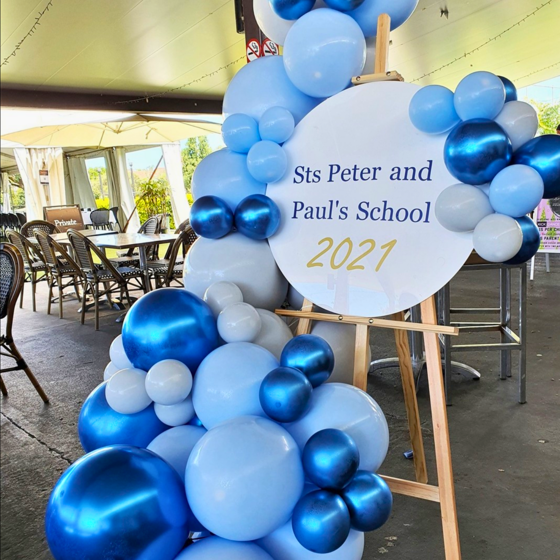 A celebratory display featuring a cluster of blue and white balloons arranged around a circular sign that reads ‘Sts Peter and Paul’s School 2021’. The arrangement is placed on an easel and is located in an outdoor setting with chairs and tables under a covered area. The festive decoration suggests a celebration or event related to the school, possibly a graduation or end-of-year ceremony for the year 2021. Prop hire, Prop hiring, Prop hire, event stylists Brisbane, Brisbane event styling