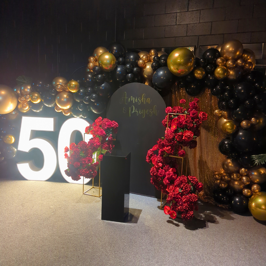 Celebration setup with a large '50' in black and gold, surrounded by a black, gold, and transparent balloon arch. Sign with 'Anisha & Rajesh' in cursive script. Red floral arrangements shaped as '5' and '0', indoors with dim lighting.  Prop hire, Prop hiring, Prop hire, event stylists Brisbane, brisbane event styling, brisbane event styling, event stylist Brisbane, Brisbane Wedding, Gold Coast Wedding, Floral Styling, Lush & Lavish Events, Brisbane Tablescape, Gold Coast Tablescapes, Brisbane Candle Hire