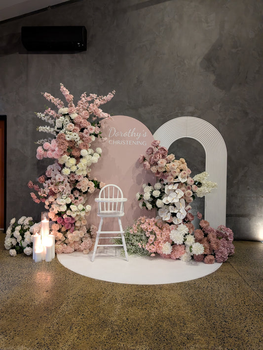 Full view of Dorothy’s christening backdrop with a blush personalised sign, white ripple arch, abundant pink and white flowers, candles, high chair and white floor mat.