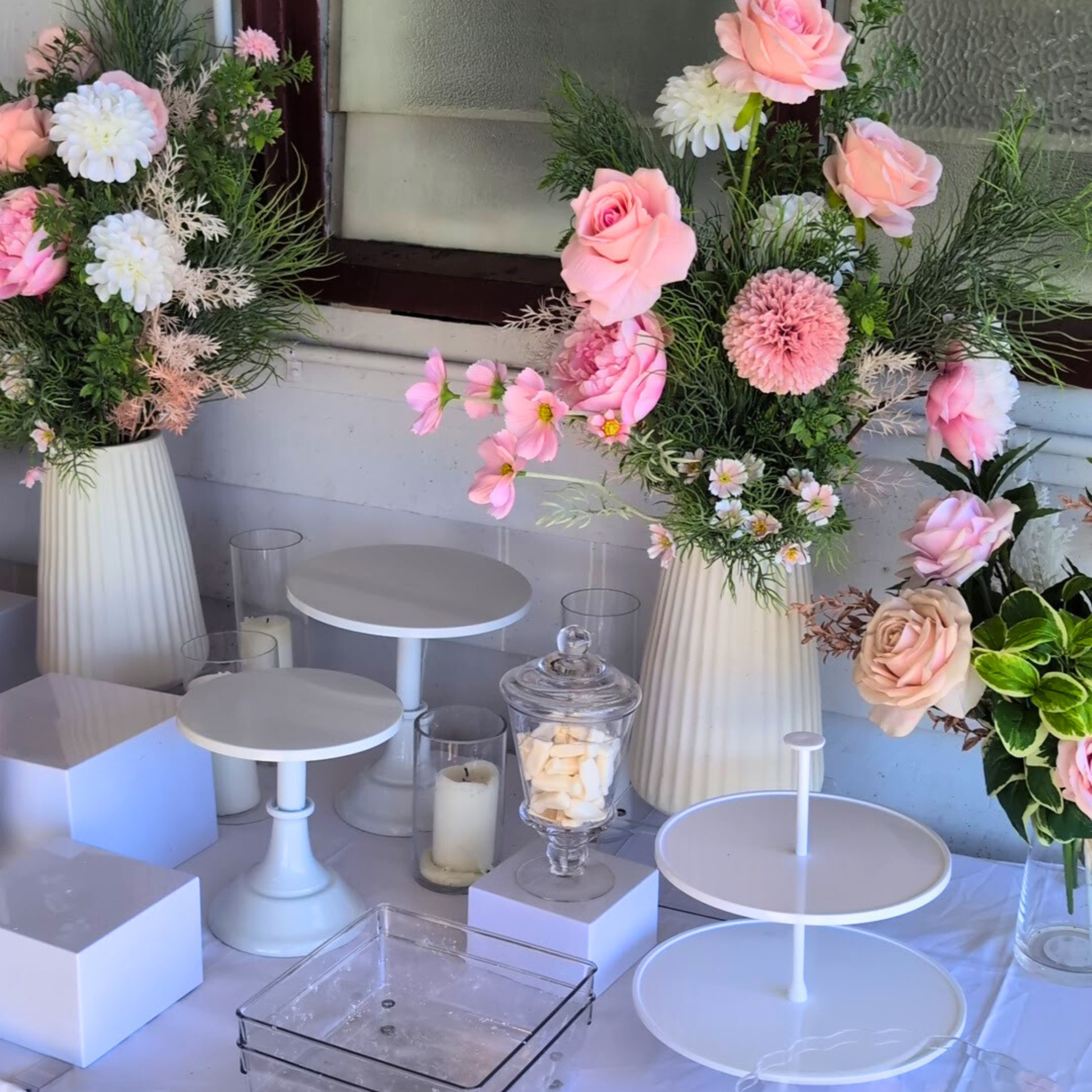 Decorative setup with floral arrangements, white stands, and candles on a table. white cake stand, cake stand set, dessert display, tiered cake stand, wedding cake stand, birthday cake stand, cupcake stand, party hire Brisbane, party hire Gold Coast, cake table display, event hire Brisbane, event hire Gold Coast, celebration tableware, elegant cake stand, white dessert stand, cake display hire, table centrepiece stand, cake presentation, party styling Brisbane, party styling Gold Coast