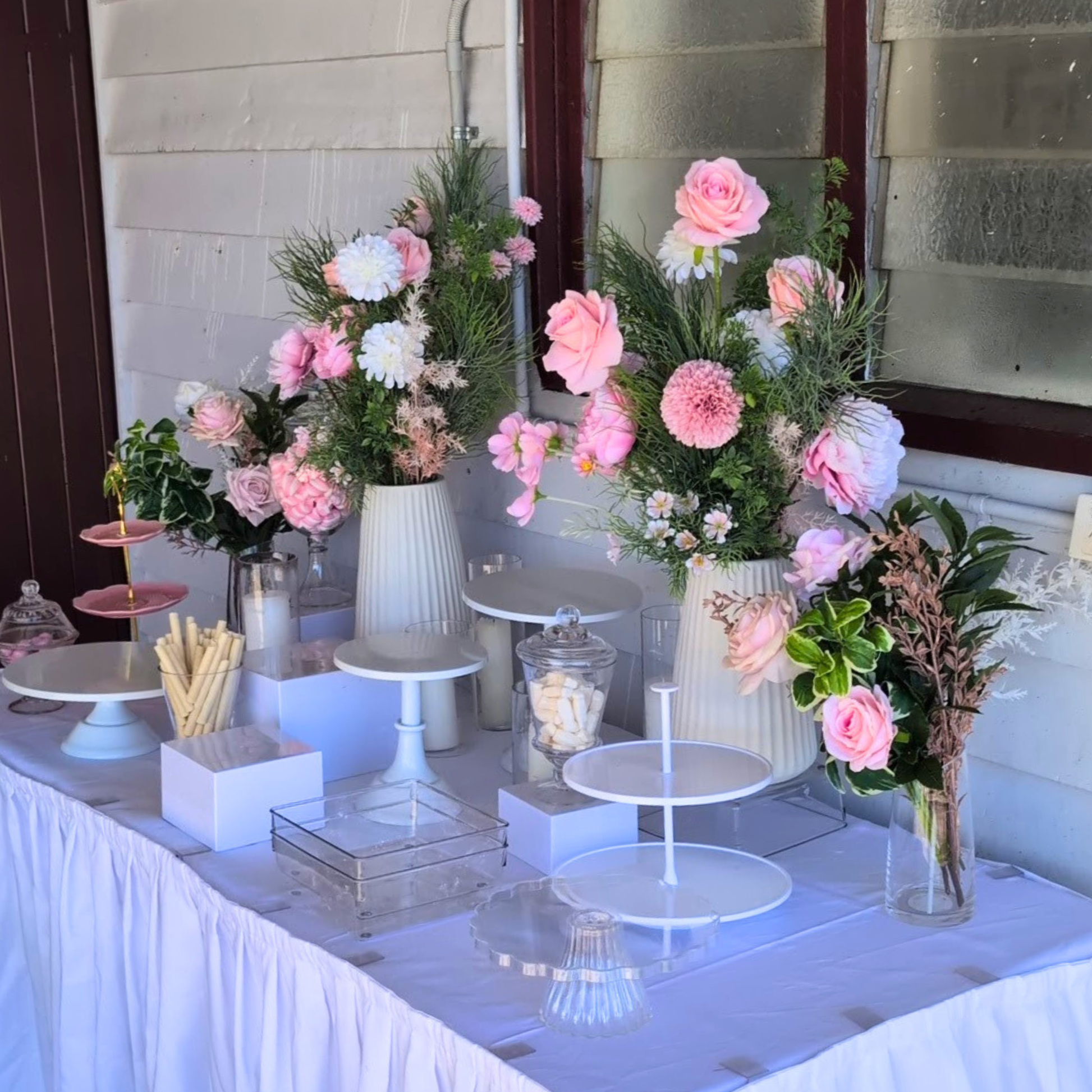 Decorative table setup with flowers, candles, and pastries on a white tablecloth. white acrylic dessert risers, dessert risers set, tiered dessert display, cupcake risers, cake table display, party hire Brisbane, party hire Gold Coast, event hire Brisbane, event hire Gold Coast, birthday dessert display, wedding dessert table, elegant dessert risers, modern dessert display, dessert table styling, sweets display stand, acrylic dessert stand, celebration tableware, party styling Brisbane, party styling Gold C