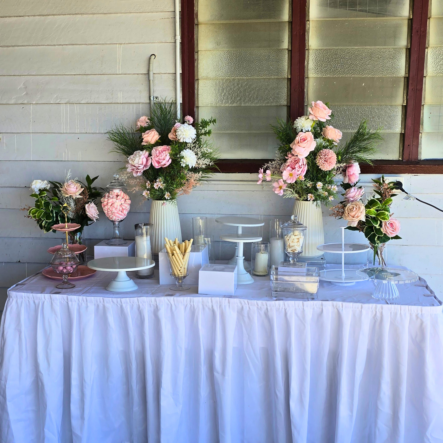 Decorative table with floral arrangements and candles against a light-colored wall. white cake stand, cake stand set, dessert display, tiered cake stand, wedding cake stand, birthday cake stand, cupcake stand, party hire Brisbane, party hire Gold Coast, cake table display, event hire Brisbane, event hire Gold Coast, celebration tableware, elegant cake stand, white dessert stand, cake display hire, table centrepiece stand, cake presentation, party styling Brisbane, party styling Gold Coast