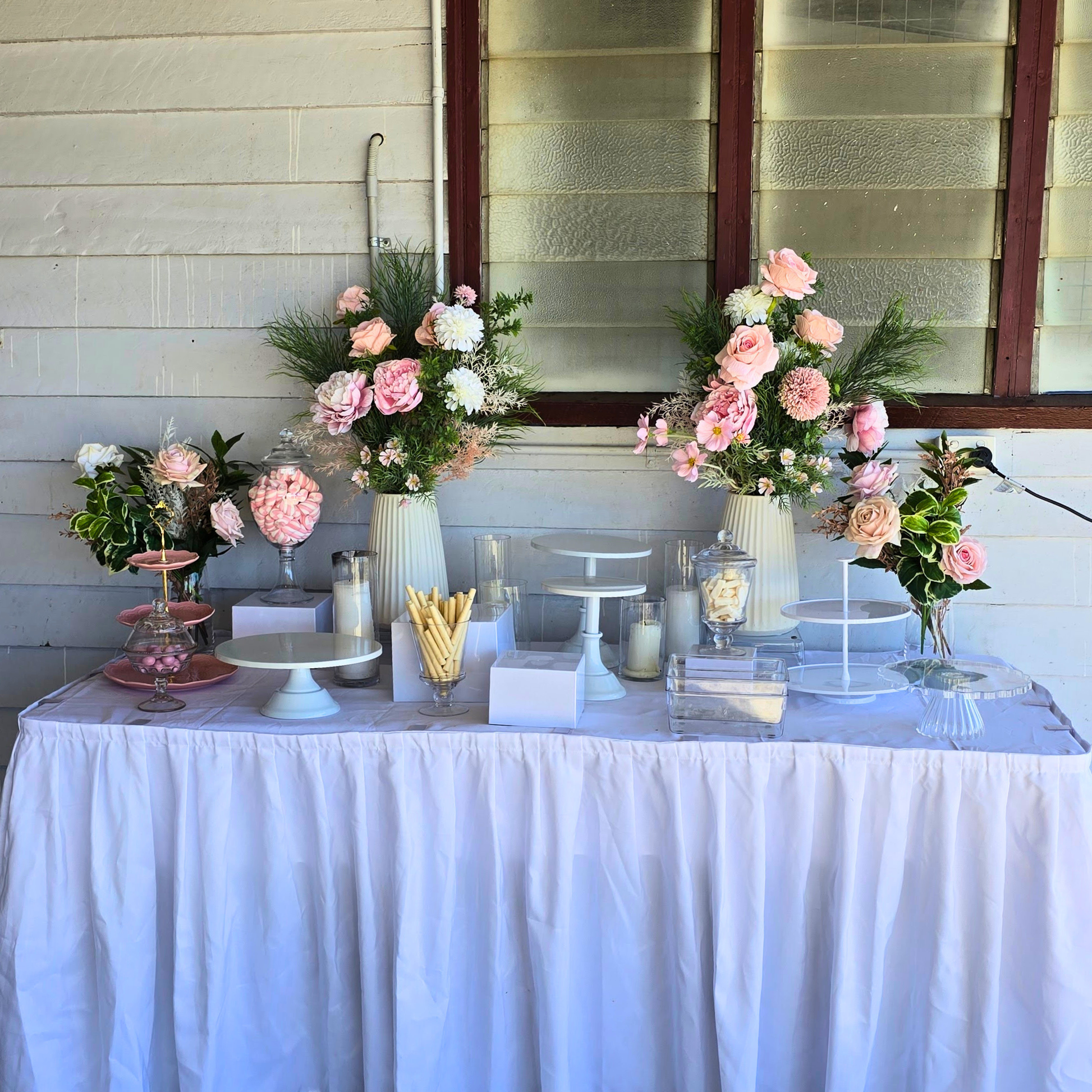 Decorative table with floral arrangements and candles against a light-colored wall. white cake stand, cake stand set, dessert display, tiered cake stand, wedding cake stand, birthday cake stand, cupcake stand, party hire Brisbane, party hire Gold Coast, cake table display, event hire Brisbane, event hire Gold Coast, celebration tableware, elegant cake stand, white dessert stand, cake display hire, table centrepiece stand, cake presentation, party styling Brisbane, party styling Gold Coast