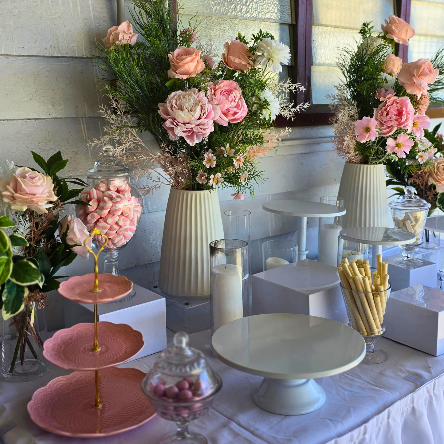 Decorative table setup with flowers, candy, and pastries in a room. white cake stand, cake stand set, dessert display, tiered cake stand, wedding cake stand, birthday cake stand, cupcake stand, party hire Brisbane, party hire Gold Coast, cake table display, event hire Brisbane, event hire Gold Coast, celebration tableware, elegant cake stand, white dessert stand, cake display hire, table centrepiece stand, cake presentation, party styling Brisbane, party styling Gold Coast