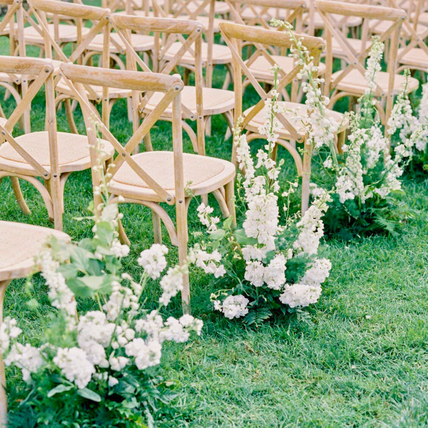 Decorative floral arrangements on grass with wooden chairs in the background. wedding aisle markers hire, floral aisle markers hire, wedding aisle flowers hire, DIY wedding aisle decor, wedding ceremony decor hire, aisle flowers for wedding, wedding flowers hire Brisbane, Brisbane wedding hire decor, wedding aisle decor Brisbane, Gold Coast wedding hire, wedding flowers hire Gold Coast, DIY wedding flowers, ceremony aisle styling, wedding aisle arrangements, modern wedding decor hire, organic wedding floral