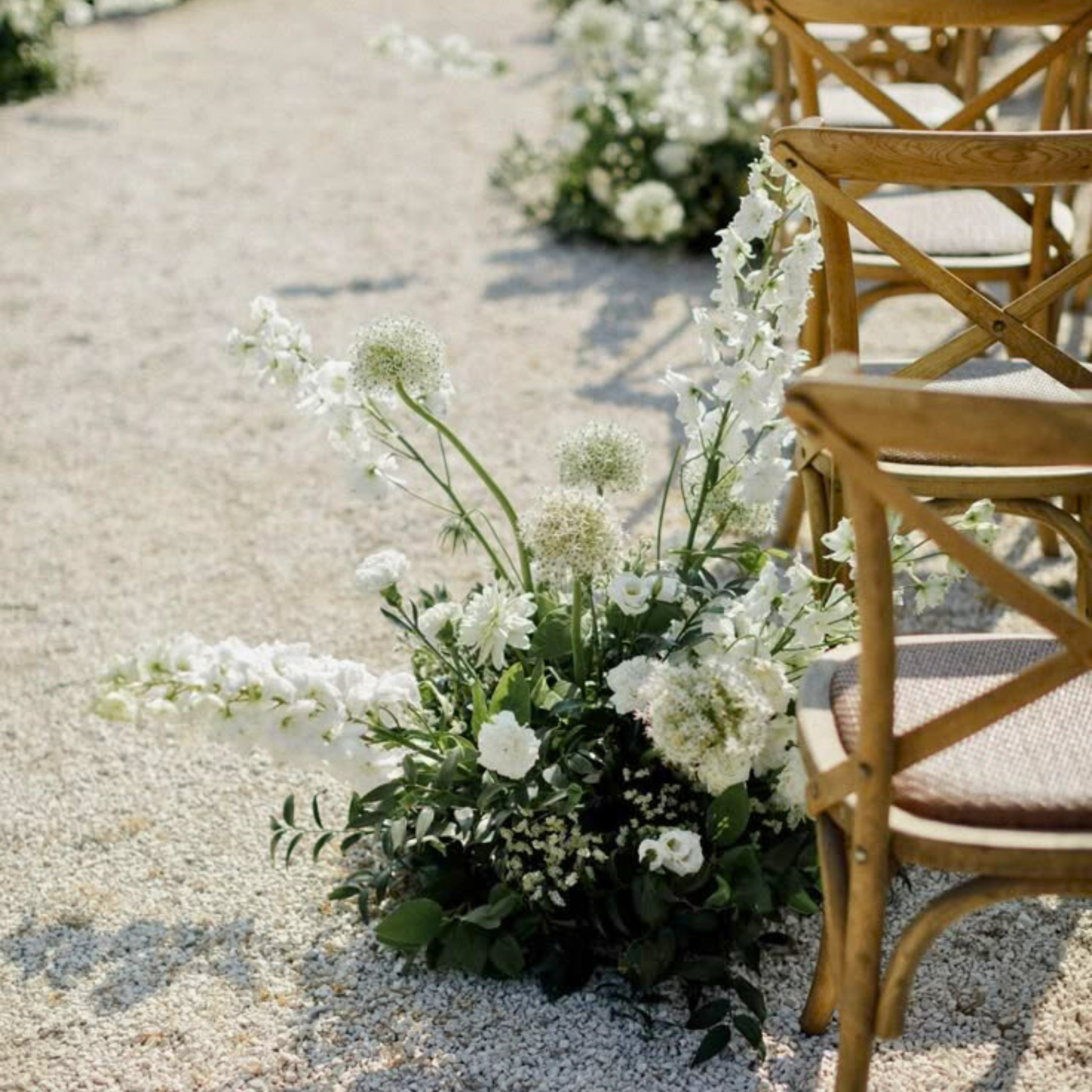 Decorative floral arrangement on a gravel path with wooden chairs in the background. wedding aisle markers hire, floral aisle markers hire, wedding aisle flowers hire, DIY wedding aisle decor, wedding ceremony decor hire, aisle flowers for wedding, wedding flowers hire Brisbane, Brisbane wedding hire decor, wedding aisle decor Brisbane, Gold Coast wedding hire, wedding flowers hire Gold Coast, DIY wedding flowers, ceremony aisle styling, wedding aisle arrangements, modern wedding decor hire, organic wedding
