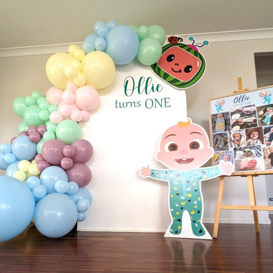 Cocomelon-themed first birthday backdrop featuring a vibrant balloon arch in blue, yellow, and pink, with a large Cocomelon character cutout and ‘Ollie turns ONE’ in elegant script. A photo collage on an easel celebrates Ollie’s first year. Prop hire, Prop hiring, Prop hire, event stylists Brisbane, brisbane event styling, brisbane event styling, event stylist Brisbane, Brisbane Wedding, Gold Coast Wedding, Floral Styling, Lush & Lavish Events