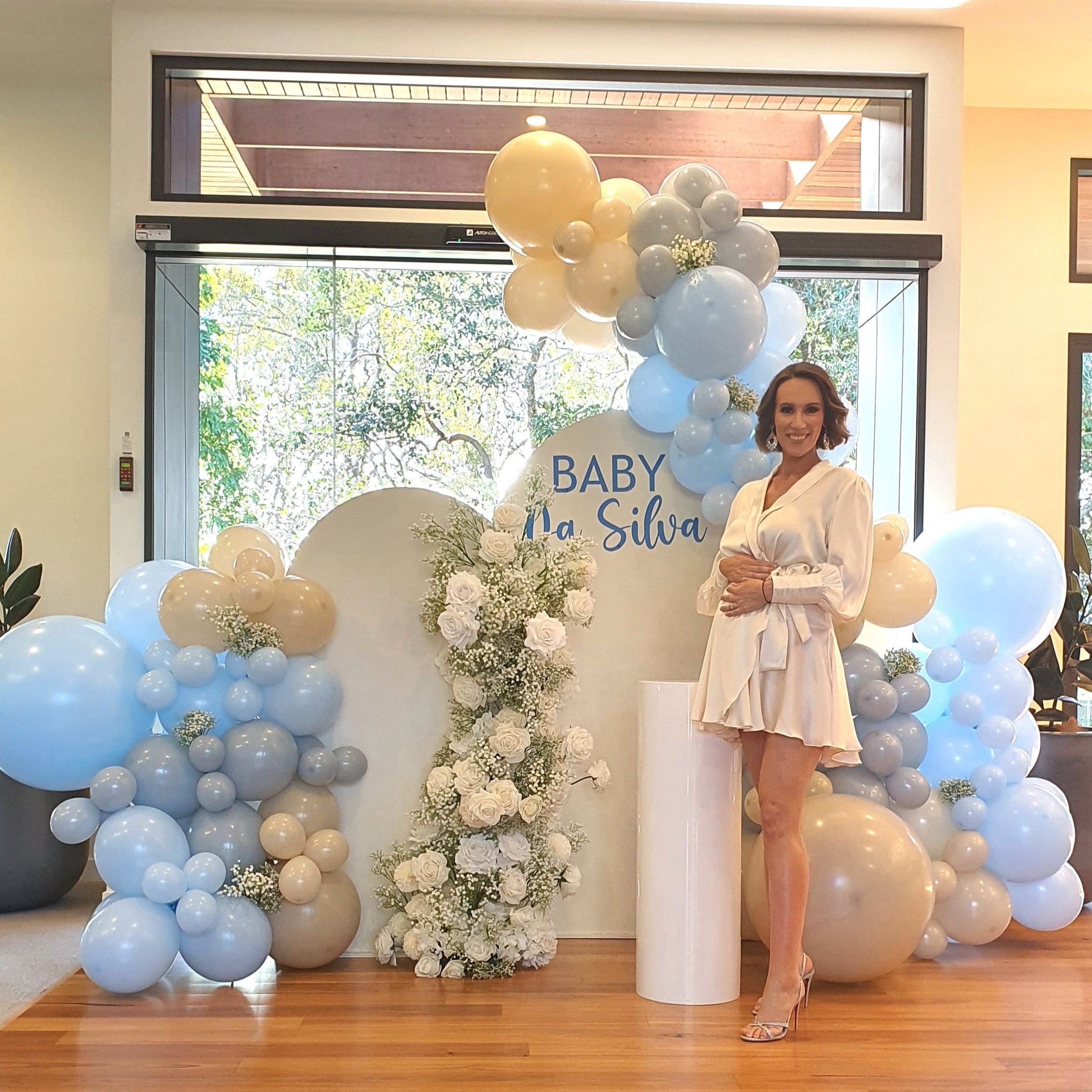 Woman in a white dress at a baby shower backdrop with blue, beige, and white balloons, and a floral arrangement. The backdrop reads "BABY Da Silva." Prop hire, Prop hiring, Prop hire, event stylists Brisbane, brisbane event styling, brisbane event styling, event stylist Brisbane, Brisbane Wedding, Gold Coast Wedding, Floral Styling, Lush & Lavish Events, Brisbane Corporate Styling, Gold Coast Corporate Styling, Brisbane Brand Activation