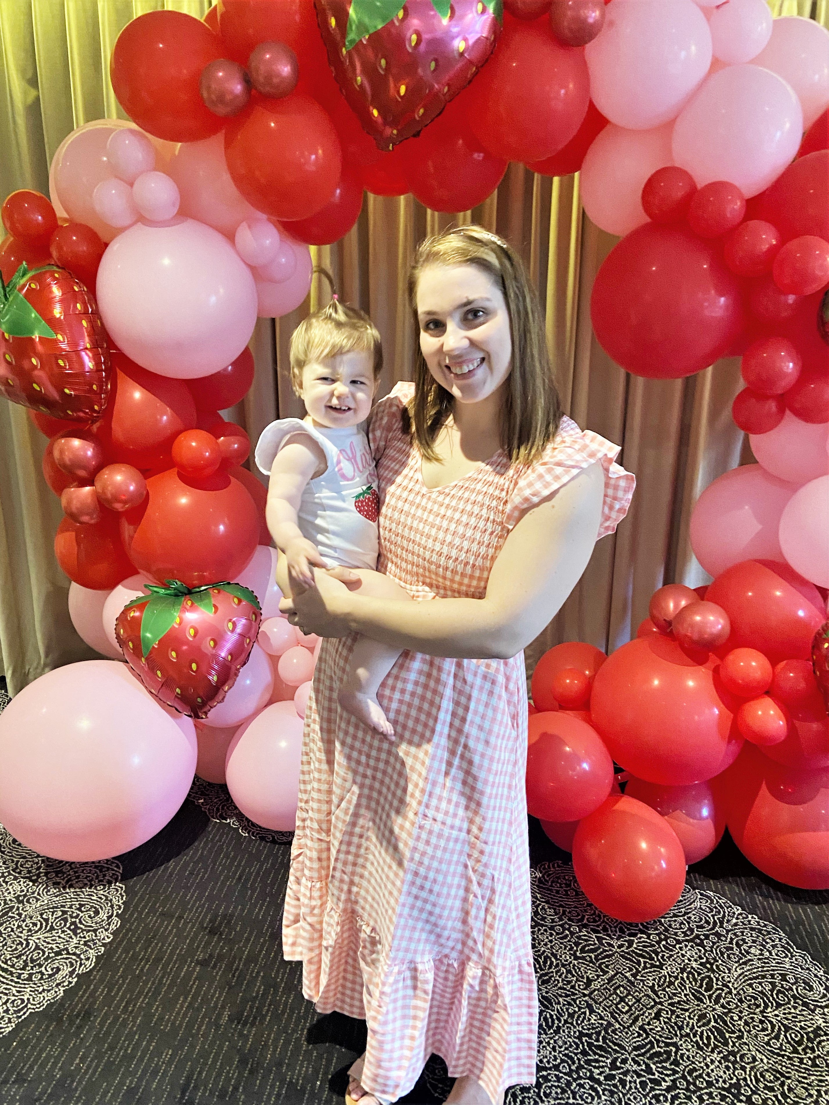 Woman holding child in front of balloon arch with red and pink balloons, including strawberry-shaped balloons. Prop hire, Prop hiring, Prop hire, event stylists Brisbane, brisbane event styling, brisbane event styling, event stylist Brisbane, Brisbane Wedding, Gold Coast Wedding, Floral Styling, Lush & Lavish Events, Brisbane Corporate Styling, Gold Coast Corporate Styling, Brisbane Brand Activation