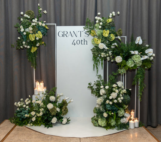 Full view of a 40th birthday backdrop with two large asymmetrical floral arrangements, personalised signage, pillar candles and a curved white floor mat