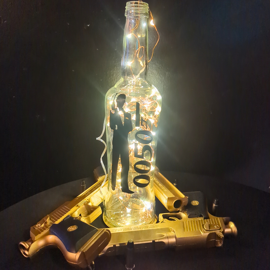 A glass bottle with illuminated light bulbs inside sits on a dark surface. Two golden toy guns form an X beneath it. Inside the bottle, a silhouette of James Bond stands next to floating numbers '0050'. The black background enhances the glow and contrast. Prop hire, Prop hiring, Prop hire, event stylists Brisbane, brisbane event styling, brisbane event styling, event stylist Brisbane, Brisbane Wedding, Gold Coast Wedding, Floral Styling, Lush & Lavish Events