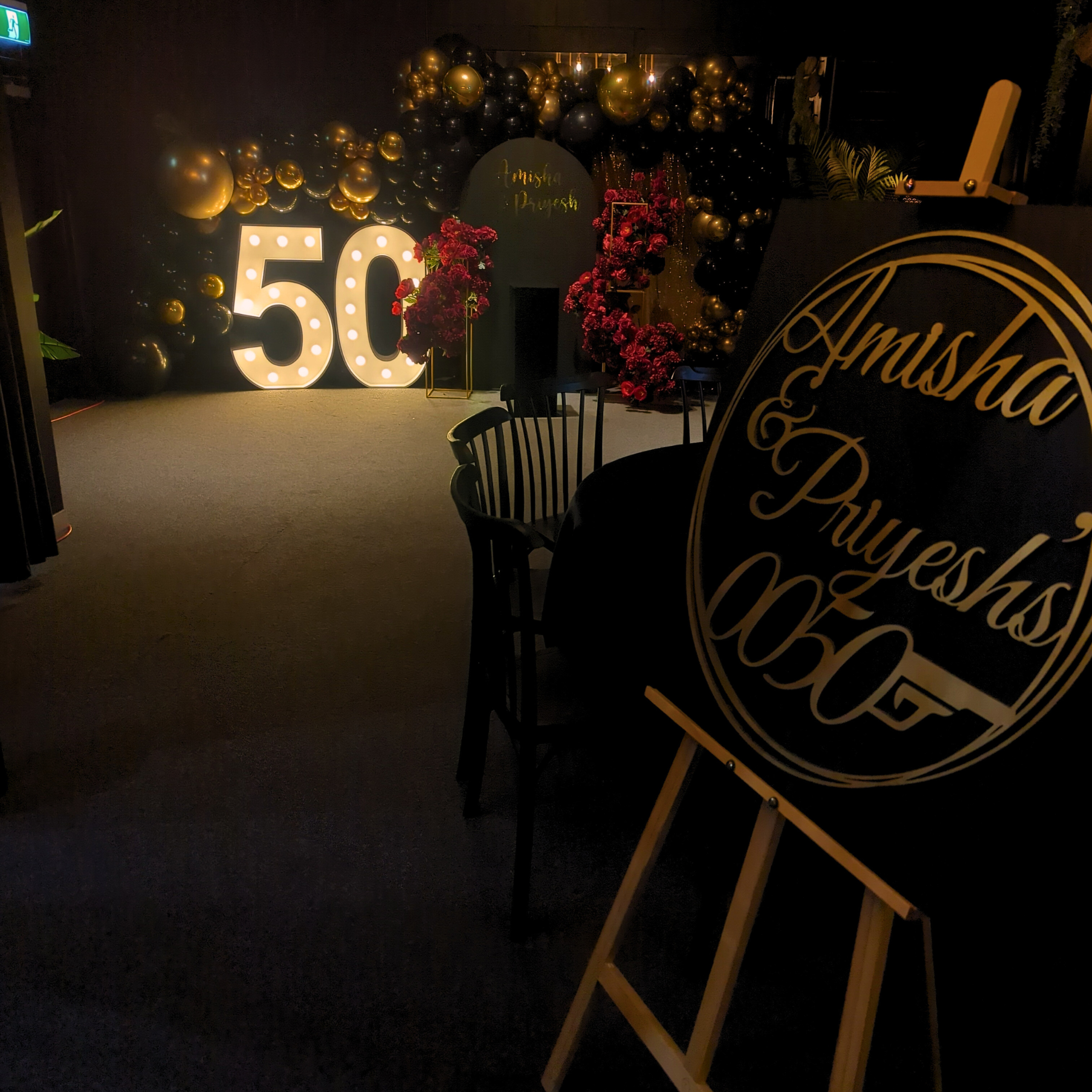 Indoor venue with dim lighting, large golden ‘50’ balloons, ‘Happy Birthday’ sign, and an easel with ‘Amisha & Priyesh’s 0050’ in elegant script. Prop hire, Prop hiring, Prop hire, event stylists Brisbane, brisbane event styling, brisbane event styling, event stylist Brisbane, Brisbane Wedding, Gold Coast Wedding, Floral Styling, Lush & Lavish Events, Brisbane Tablescape, Gold Coast Tablescapes, Brisbane Candle Hire