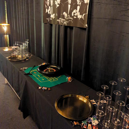 A casino-themed dessert table with a green roulette layout, chips, dice, playing cards, a tray, and a champagne glass pyramid. The background features a dark curtain with images of movie stars playing poker, all under dim lighting. Prop hire, Prop hiring, Prop hire, event stylists Brisbane, brisbane event styling, brisbane event styling, event stylist Brisbane, Brisbane Wedding, Gold Coast Wedding, Floral Styling, Lush & Lavish Events