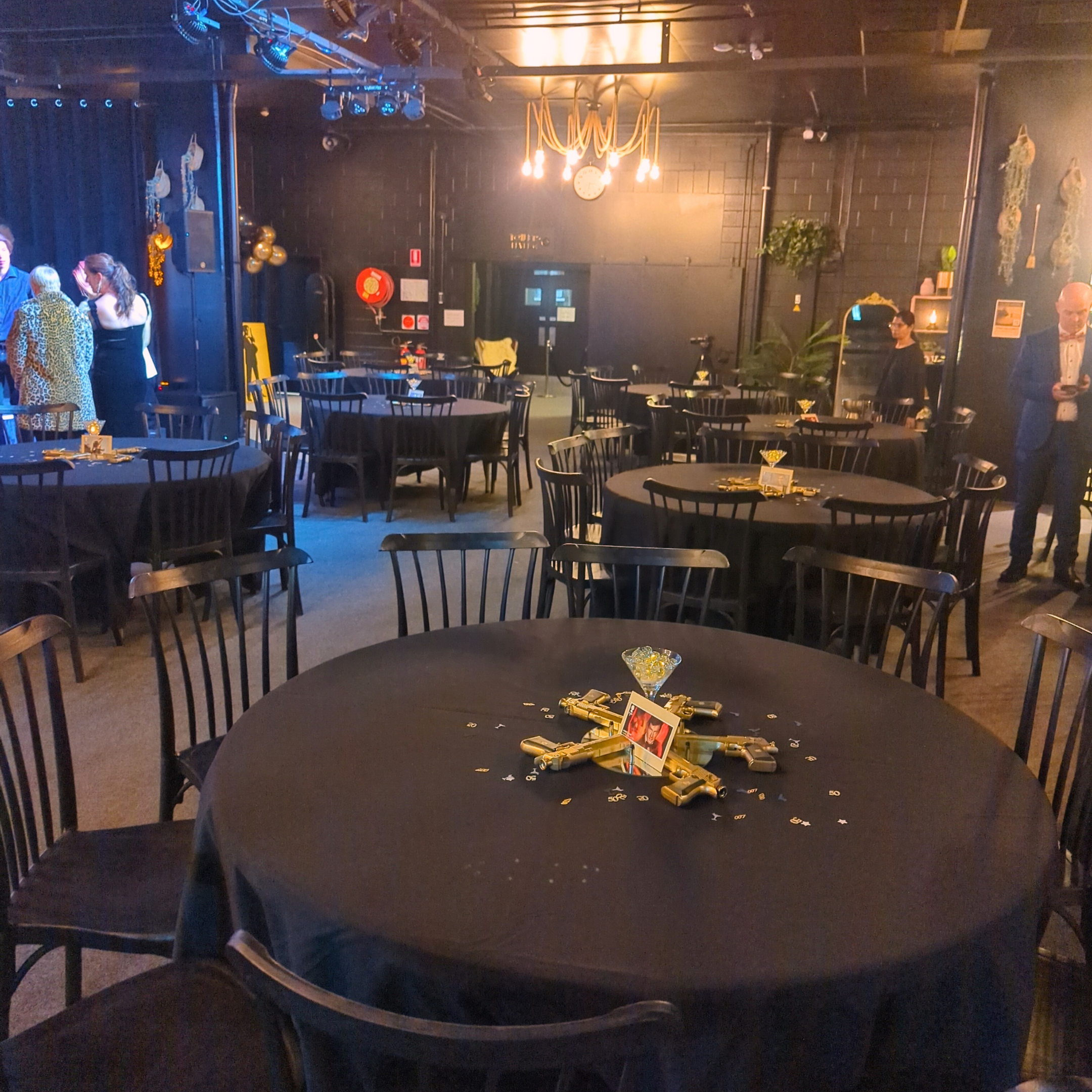 Indoor event space with black tablecloths, a table with confetti, gold guns, and custom centerpieces. Guests mingle near a bar under warm lighting. Prop hire, Prop hiring, Prop hire, event stylists Brisbane, brisbane event styling, brisbane event styling, event stylist Brisbane, Brisbane Wedding, Gold Coast Wedding, Floral Styling, Lush & Lavish Events