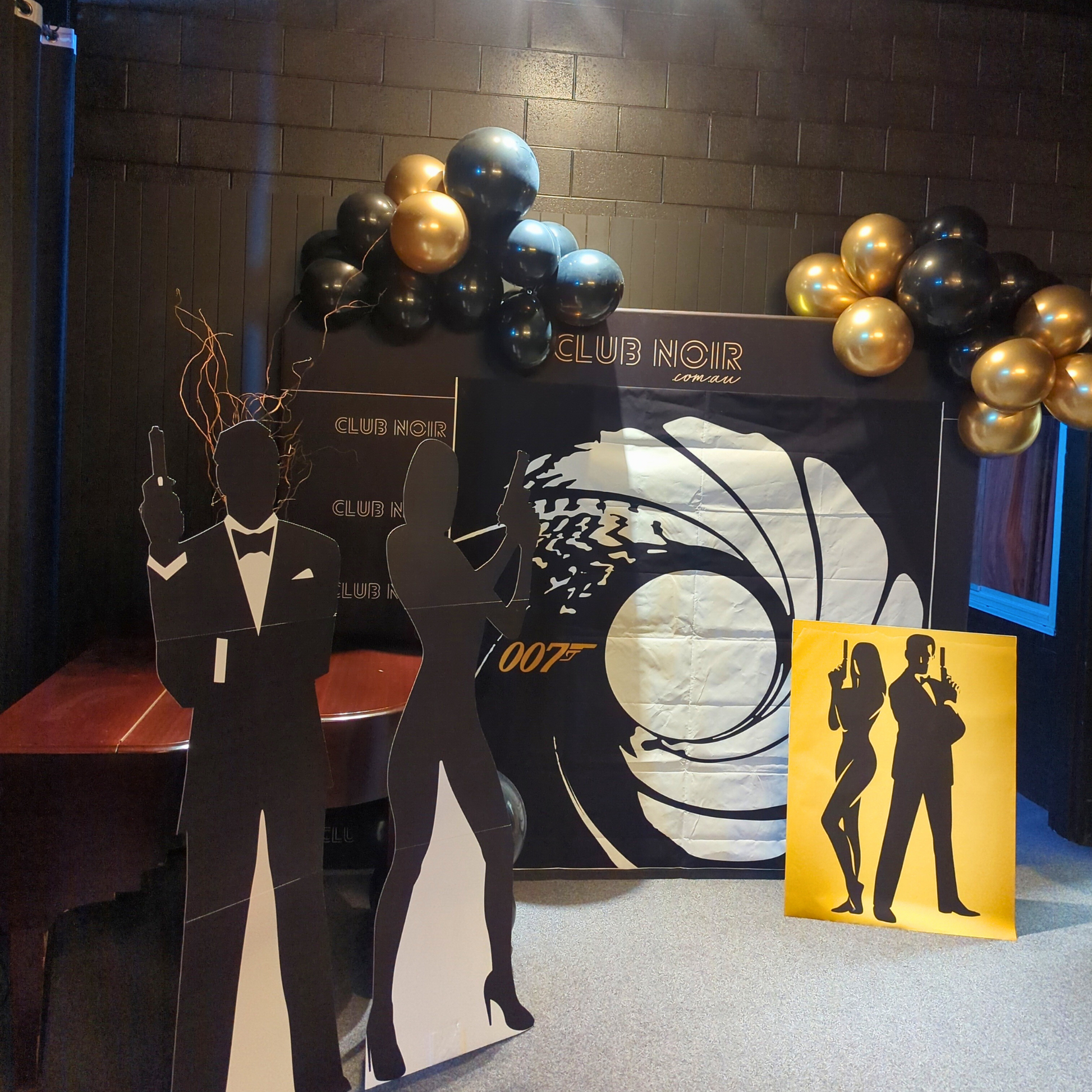 'Club Noir' themed party setup with '007' backdrop, black and gold balloons, and silhouettes of a man and woman holding guns. Prop hire, Prop hiring, Prop hire, event stylists Brisbane, brisbane event styling, brisbane event styling, event stylist Brisbane, Brisbane Wedding, Gold Coast Wedding, Floral Styling, Lush & Lavish Events, Brisbane Tablescape, Gold Coast Tablescapes, Brisbane Candle Hire