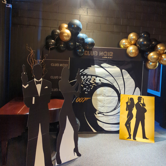 'Club Noir' themed party setup with '007' backdrop, black and gold balloons, and silhouettes of a man and woman holding guns. Prop hire, Prop hiring, Prop hire, event stylists Brisbane, brisbane event styling, brisbane event styling, event stylist Brisbane, Brisbane Wedding, Gold Coast Wedding, Floral Styling, Lush & Lavish Events, Brisbane Tablescape, Gold Coast Tablescapes, Brisbane Candle Hire