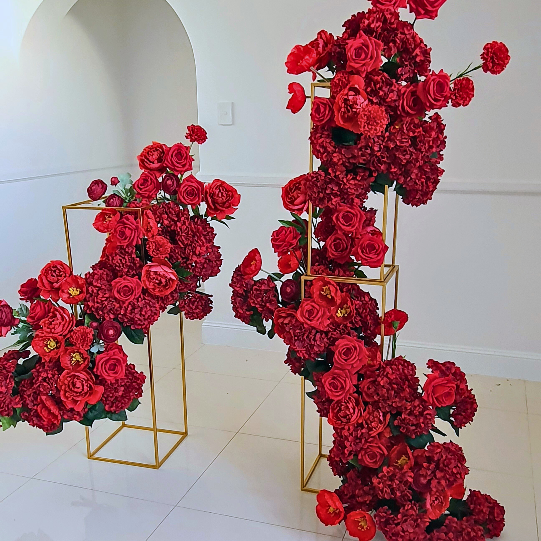 Red roses and burgundy peonies densely packed within two interlocking golden cubes on a white floor, set against a white wall with an arched doorway, creating a striking contrast. Prop hire, Prop hiring, Prop hire, event stylists Brisbane, brisbane event styling, brisbane event styling, event stylist Brisbane, Brisbane Wedding, Gold Coast Wedding, Floral Styling, Lush & Lavish Events