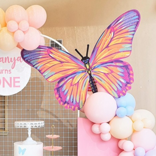Vibrant butterfly decoration with purple, pink, yellow, and blue wings perched on a pastel balloon arrangement at an outdoor event. Background features a dessert table with a white cake stand. Prop hire, Prop hiring, Prop hire, event stylists Brisbane, brisbane event styling, brisbane event styling, event stylist Brisbane, Brisbane Wedding, Gold Coast Wedding, Floral Styling, Lush & Lavish Events