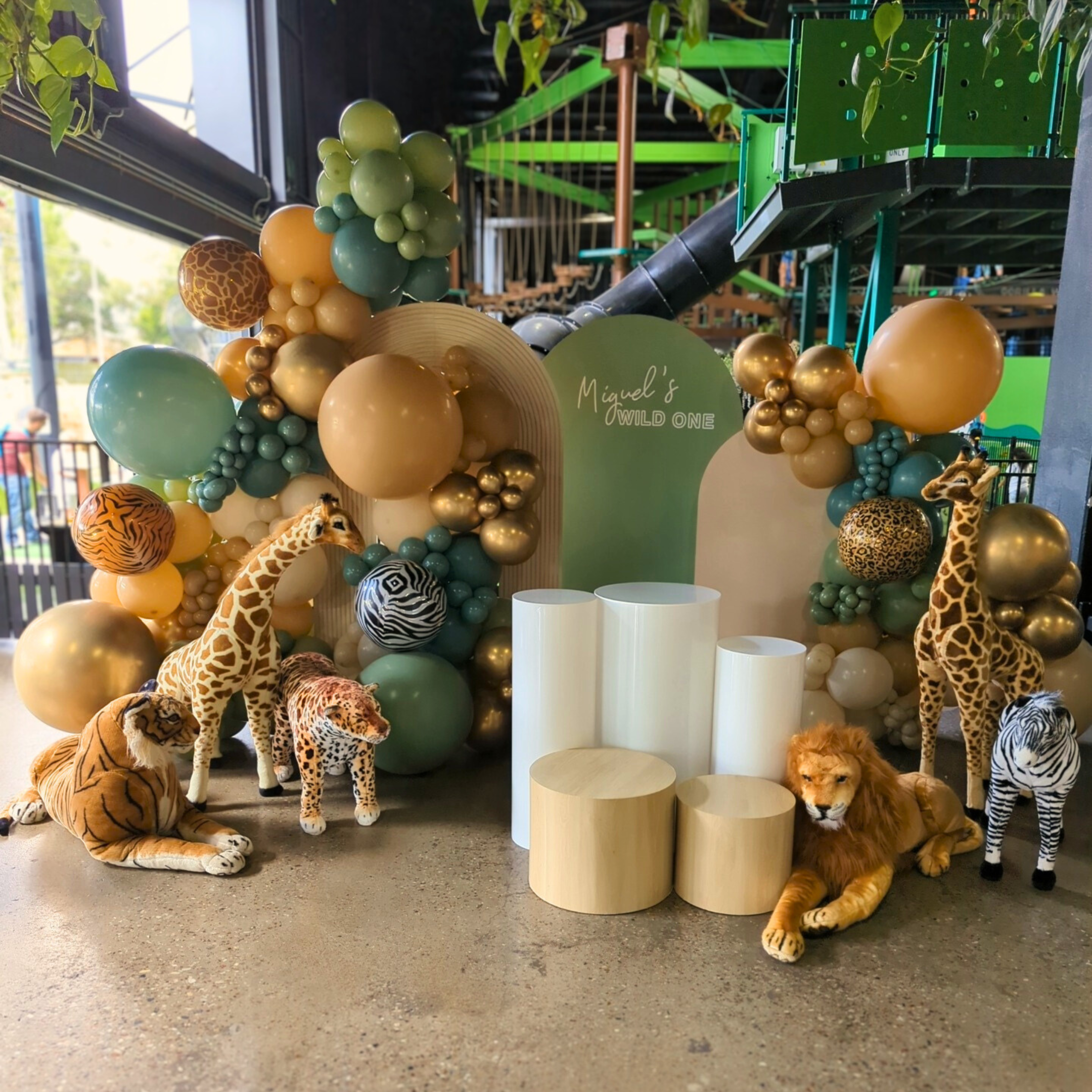 Jungle-themed backdrop with "Miguel's Wild One" sign and green, gold, and orange balloons, plush animals, and white stands. Prop hire, Prop hiring, Prop hire, event stylists Brisbane, brisbane event styling, brisbane event styling, event stylist Brisbane, Brisbane Wedding, Gold Coast Wedding, Floral Styling, Lush & Lavish Events, Brisbane Corporate Styling, Gold Coast Corporate Styling, Brisbane Brand Activation