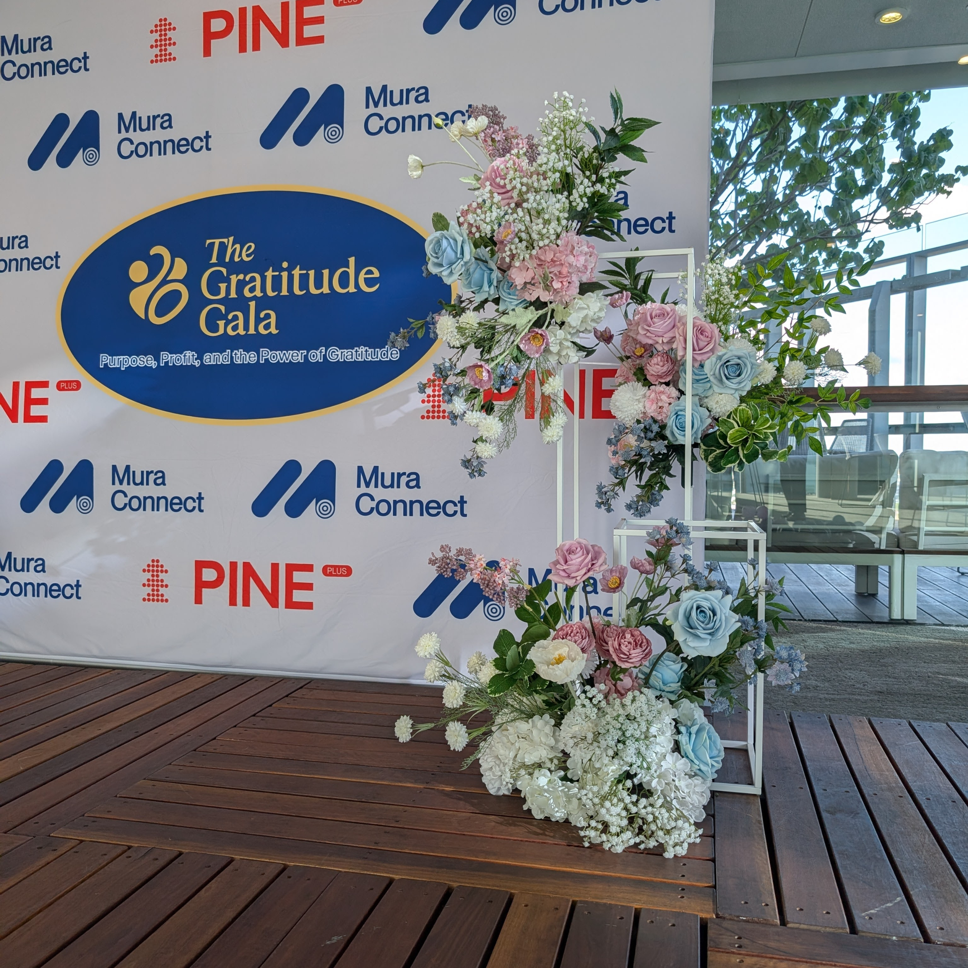 Floral arrangement in front of a backdrop with 'The Gratitude Gala' and brand logos. media wall balloon styling, balloon garland installation, corporate balloon styling, brand activation balloons, media wall styling service, campaign balloon install, branded event styling, product launch balloon decor, PR event styling balloons, corporate event decor hire, balloon installation service, event styling brisbane, balloon styling brisbane, corporate events brisbane, brand activation brisbane, event styling gold 