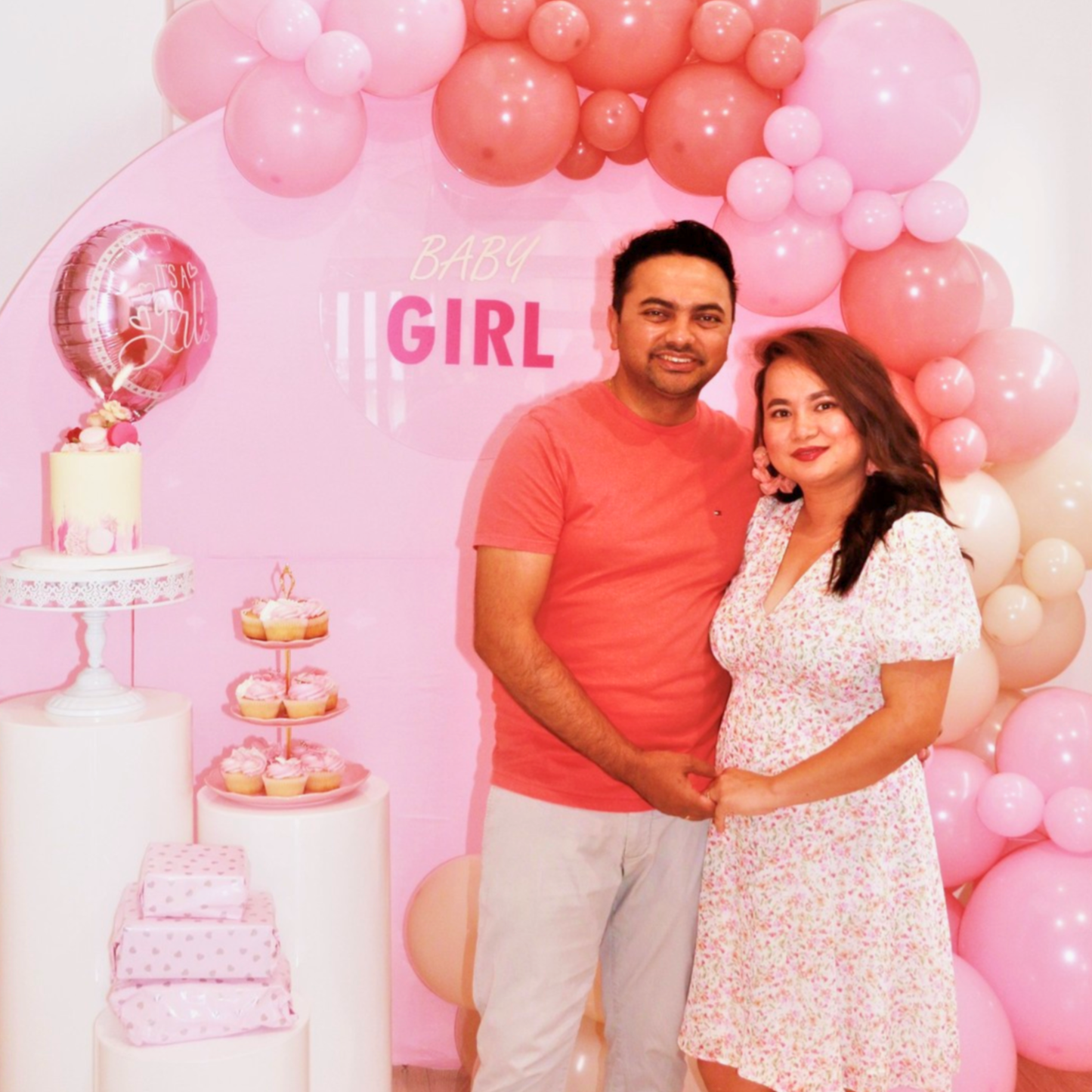 Pregnant couple posing in front of pink balloons and 'It's a Girl' sign with a cake and decorations. pink cupcake stand hire, blue cupcake stand hire, 3 tier cupcake stand hire, cupcake stand hire brisbane, cupcake stand hire gold coast, kids party cupcake stand, birthday party dessert stand, baby shower cupcake stand, party decor hire qld, dessert table styling hire, colourful cupcake stand, party prop hire brisbane, party hire gold coast, kids birthday party decor, cupcake display stand hire, themed party