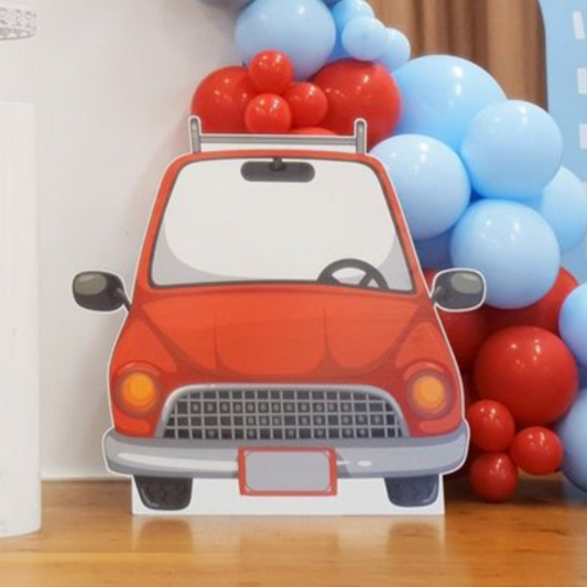 Red cartoon car cutout with big round headlights and mirrors in front of a blue and red balloon backdrop, perfect for a children’s party or automobile-themed celebration. Lush and lavish events