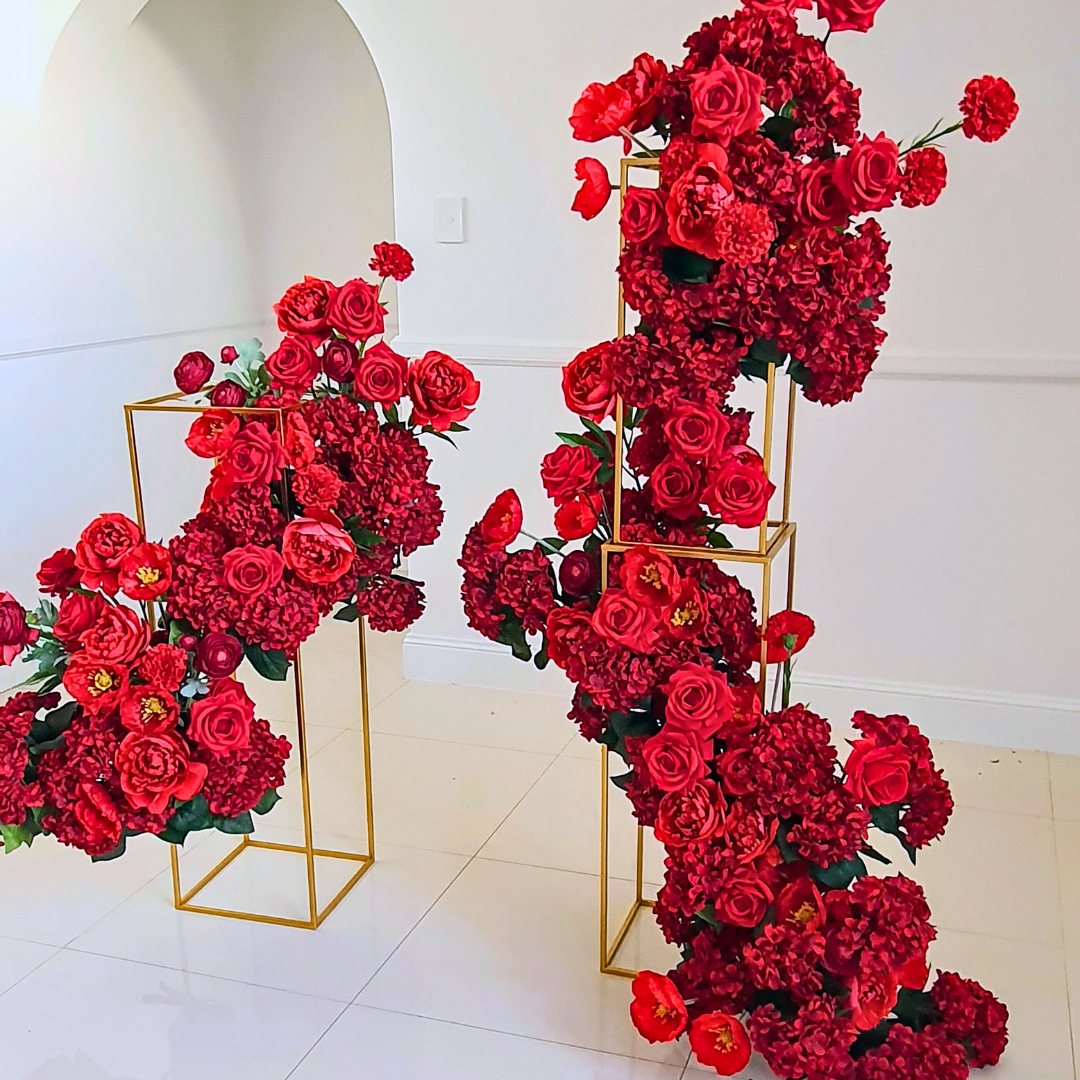 Two geometric gold stands with red flowers, predominantly roses and hydrangeas, on a white floor against a white wall with an arched doorway. Prop hire, Prop hiring, Prop hire, event stylists Brisbane, brisbane event styling, brisbane event styling, event stylist Brisbane, Brisbane Wedding, Gold Coast Wedding, Floral Styling, Lush & Lavish Events, Brisbane Tablescape, Gold Coast Tablescapes, Brisbane Candle Hire