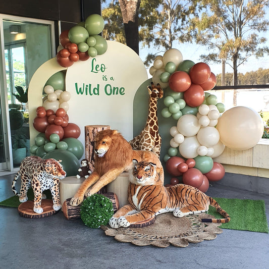 A safari-themed birthday party backdrop with a large balloon arch in shades of green, brown, and beige. The arch frames a sign that says "Leo is a Wild One." In front of the arch are stuffed animals of a lion, giraffe, tiger, and leopard. Prop hire, Prop hiring, Prop hire, event stylists Brisbane, brisbane event styling, brisbane event styling, event stylist Brisbane, Brisbane Wedding, Gold Coast Wedding, Floral Styling, Lush & Lavish Events, Brisbane Tablescape, Gold Coast Tablescapes, Brisbane Candle Hire