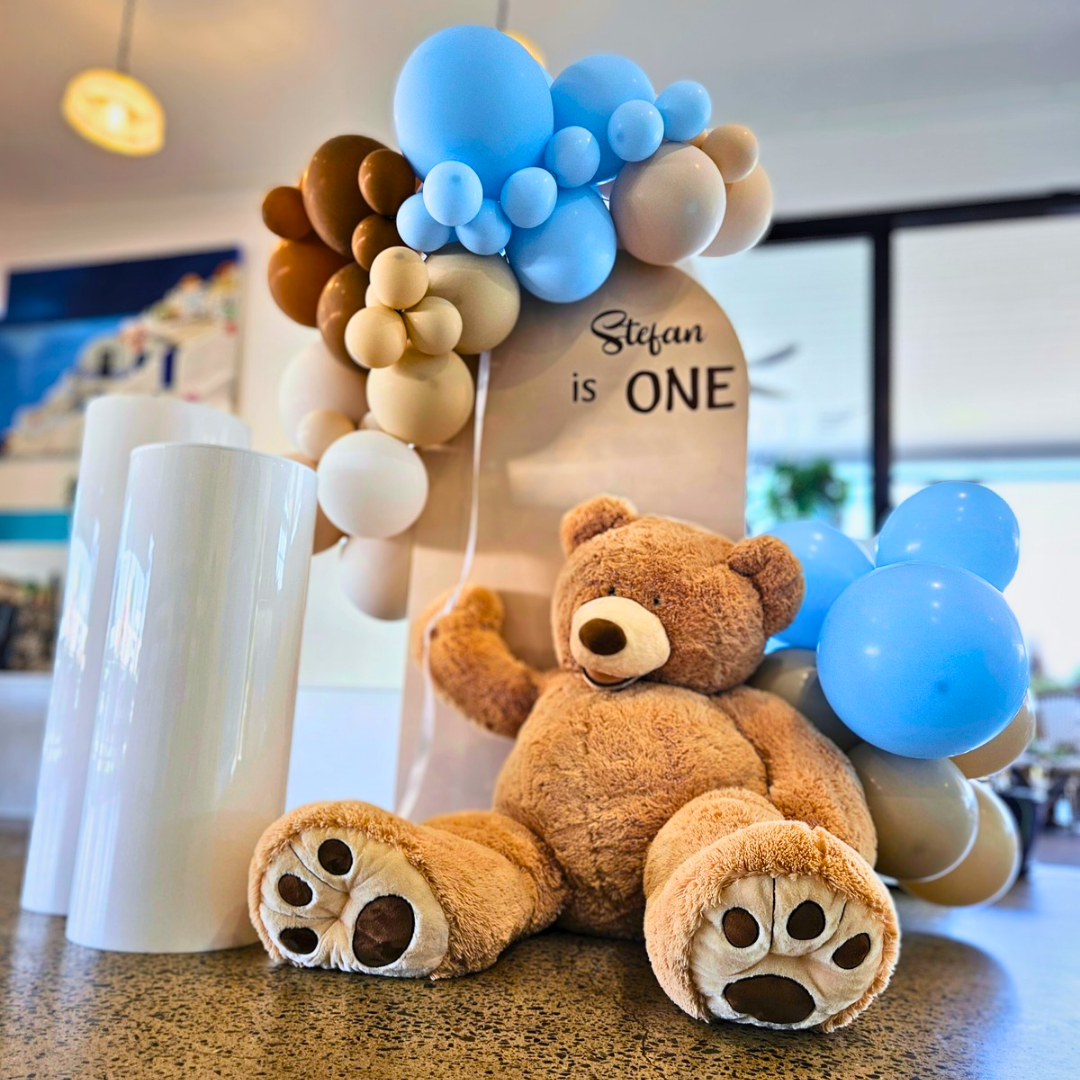 balloons backdrop is neutrals and blues with a teddy bear holding a cluster of balloons. Signage says "Stefan is ONE". Prop hire, Prop hiring, Prop hire, event stylists Brisbane, brisbane event styling, brisbane event styling, event stylist Brisbane, Brisbane Wedding, Gold Coast Wedding, Floral Styling, Lush & Lavish Events, Brisbane Corporate Styling, Gold Coast Corporate Styling, Brisbane Brand Activation, Gold Coast Tablescapes, Brisbane Candle Hire